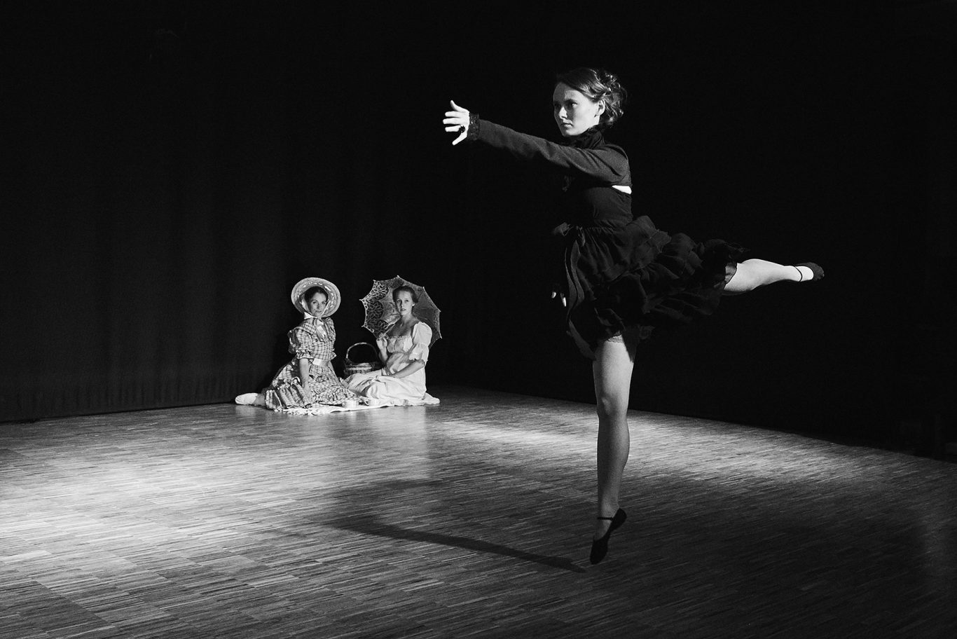 Schwarzweißfoto: Balletttänzerin in Spitzenschuhen beim arabesque auf Bühne, zwei sitzende Kinder im Hintergrund.