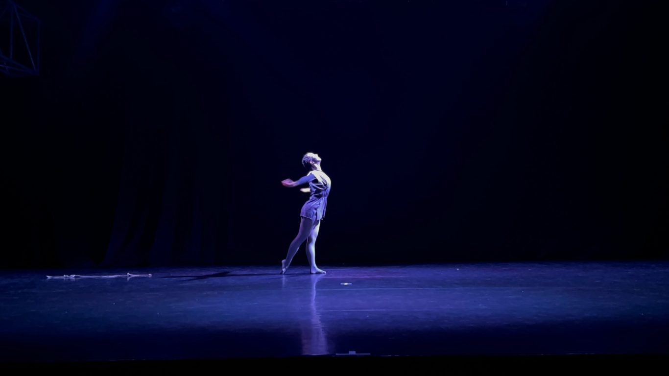 Einzelne Person in heller Kleidung tanzt auf dunkler Bühne mit blauem Scheinlicht.