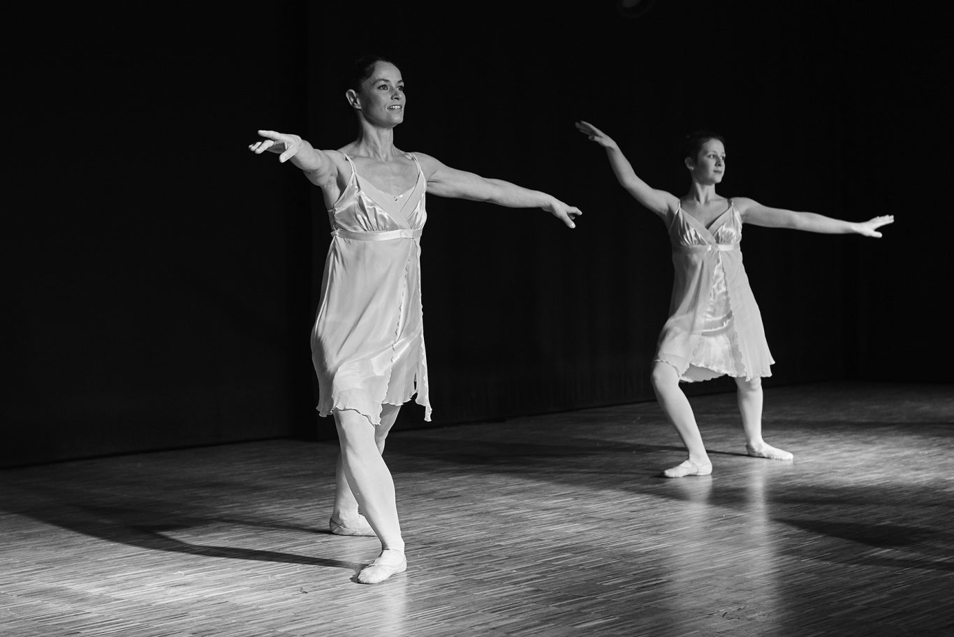 Zwei Ballerinas in Tutu und Spitzenschuhen tanzen auf einer dunklen Bühne.