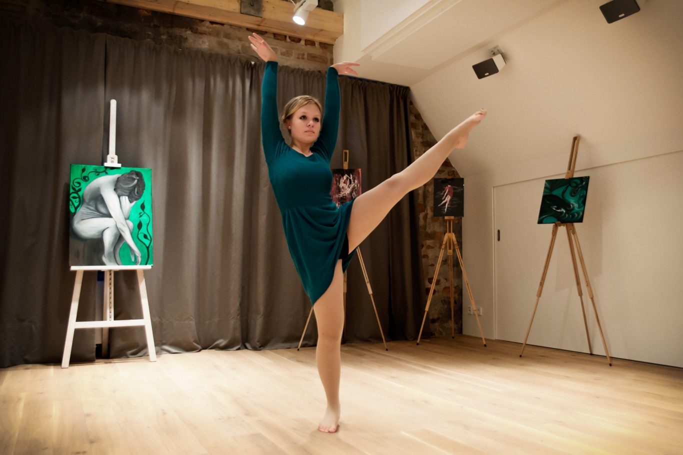 Balletttänzerin in grünem Kostüm auf Holzparkett, ein Bein gehoben, Atelierelemente im Hintergrund.