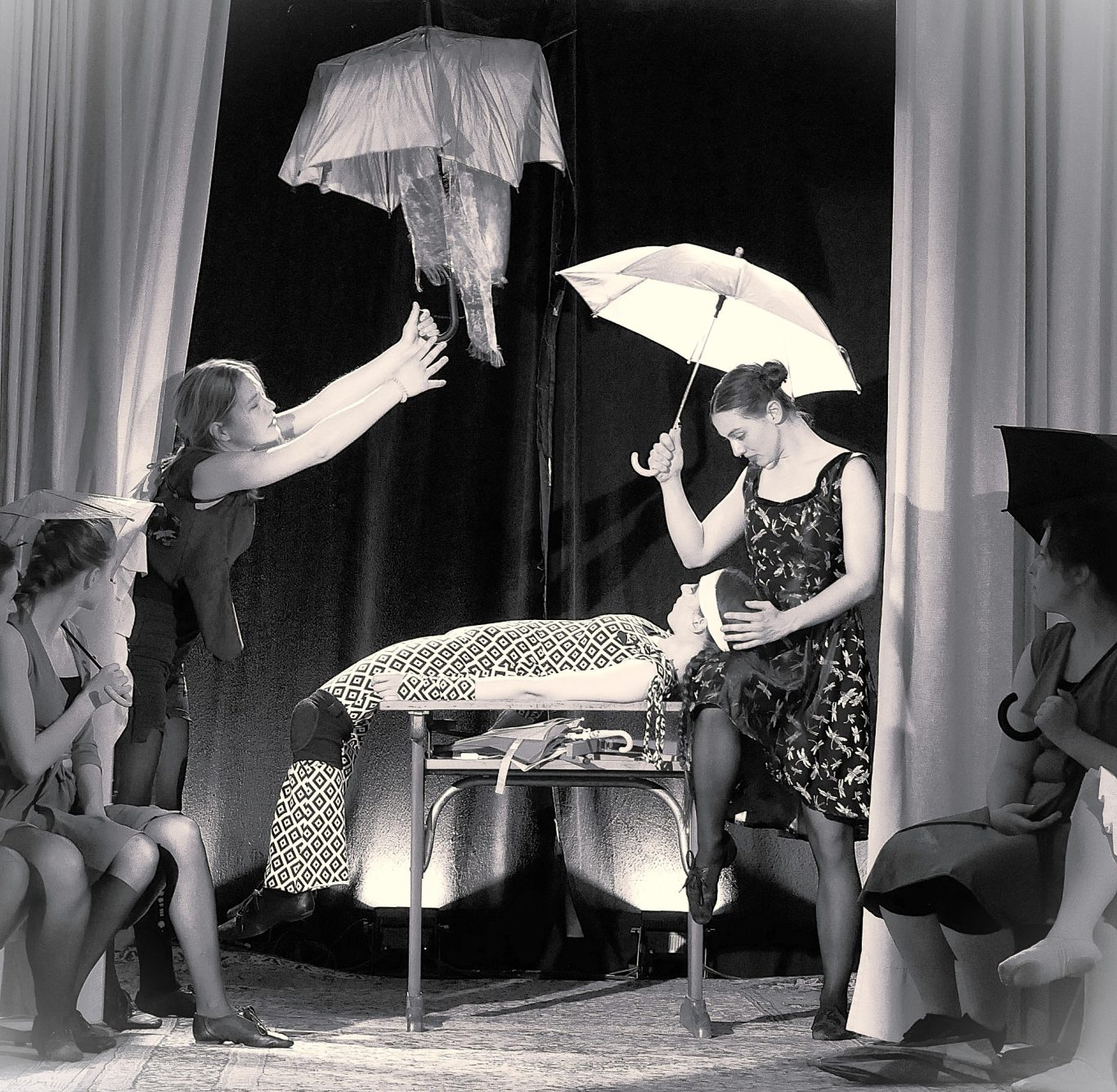 Drei Frauen sitzen um einen Tisch; zwei halten Regenschirme, eine steht mit geöffnetem Schirm über dem Tisch.