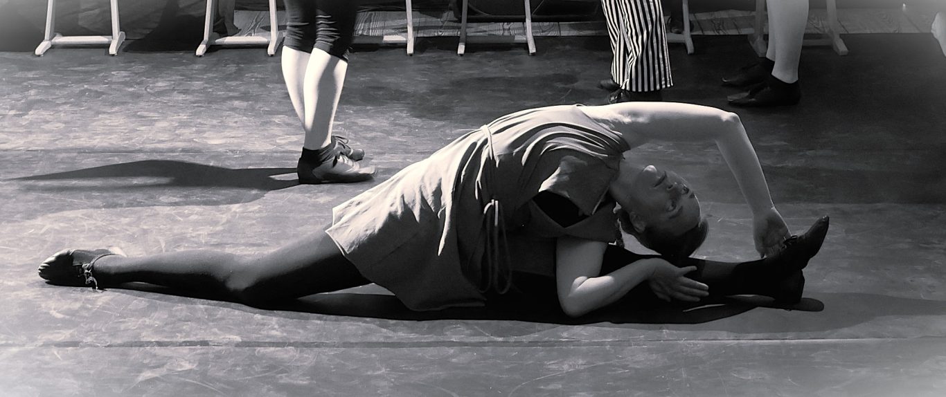 Schwarzweißfoto: Balletttänzerin in Bodendehnung, ein Bein gestreckt, Kopf auf Arm ruhend.