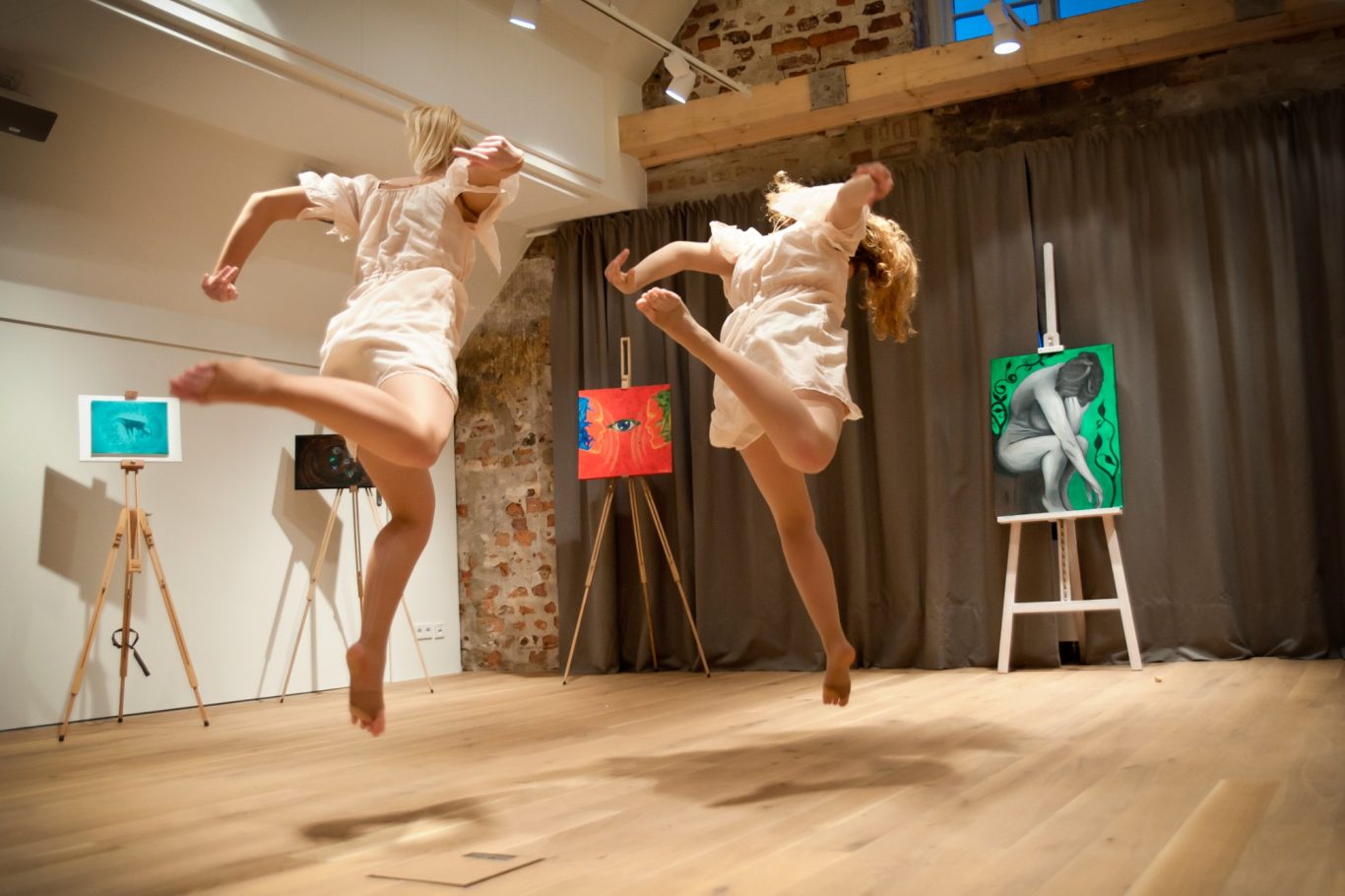 Zwei Balletttänzerinnen in Bewegung auf Holzboden vor Staffeleien mit bunten Gemälden.