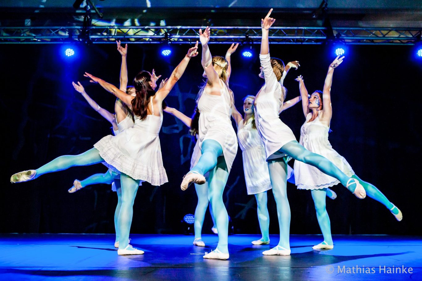 Sieben Balletttänzerinnen in weißen Tutus, auf Zehenspitzen, in synchroner Sprungpose auf Bühne.