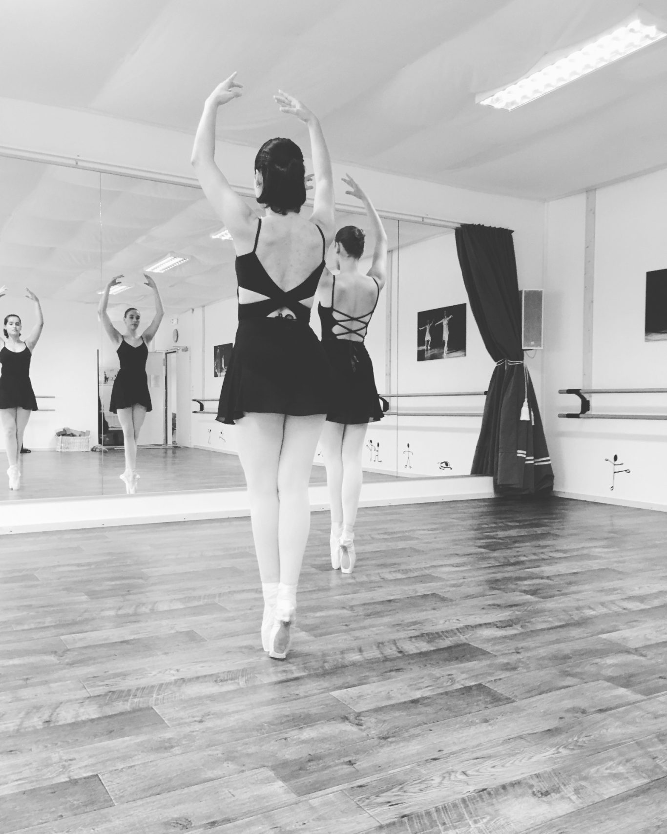 Balletttänzerin von hinten im Tanzstudio, Arme erhoben, Spiegelwand im Hintergrund.