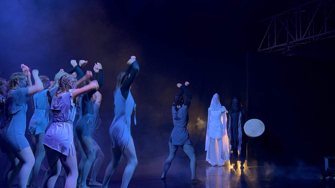 Theaterbühne mit Tänzern in bunten Kostümen unter blauer Bühnebeleuchtung.