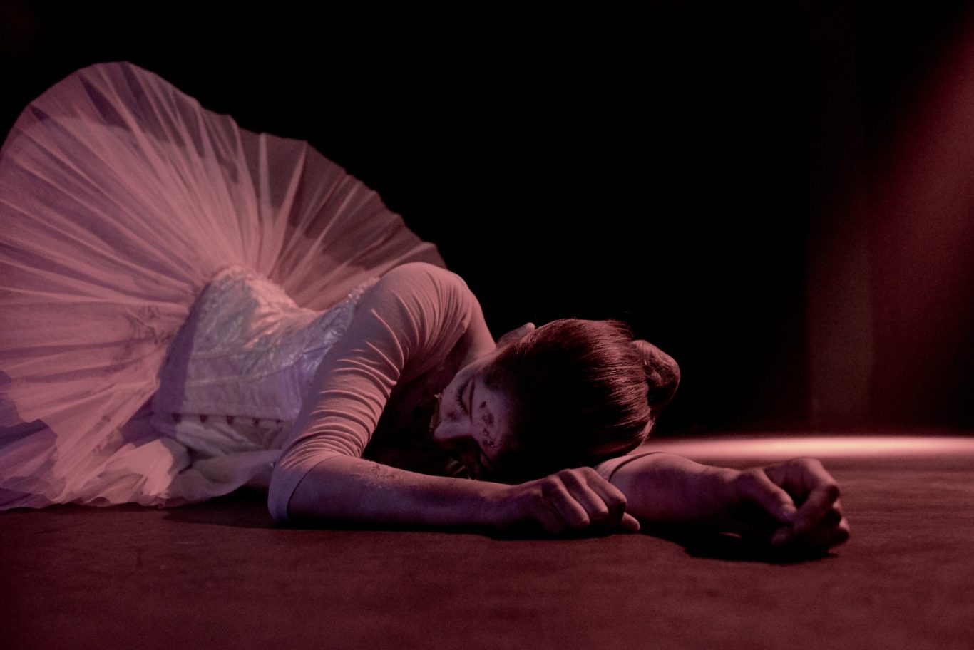 Ballerina in weißem Tutu liegt seitlich am Bühnenboden, Kopf auf ausgestrecktem Arm.