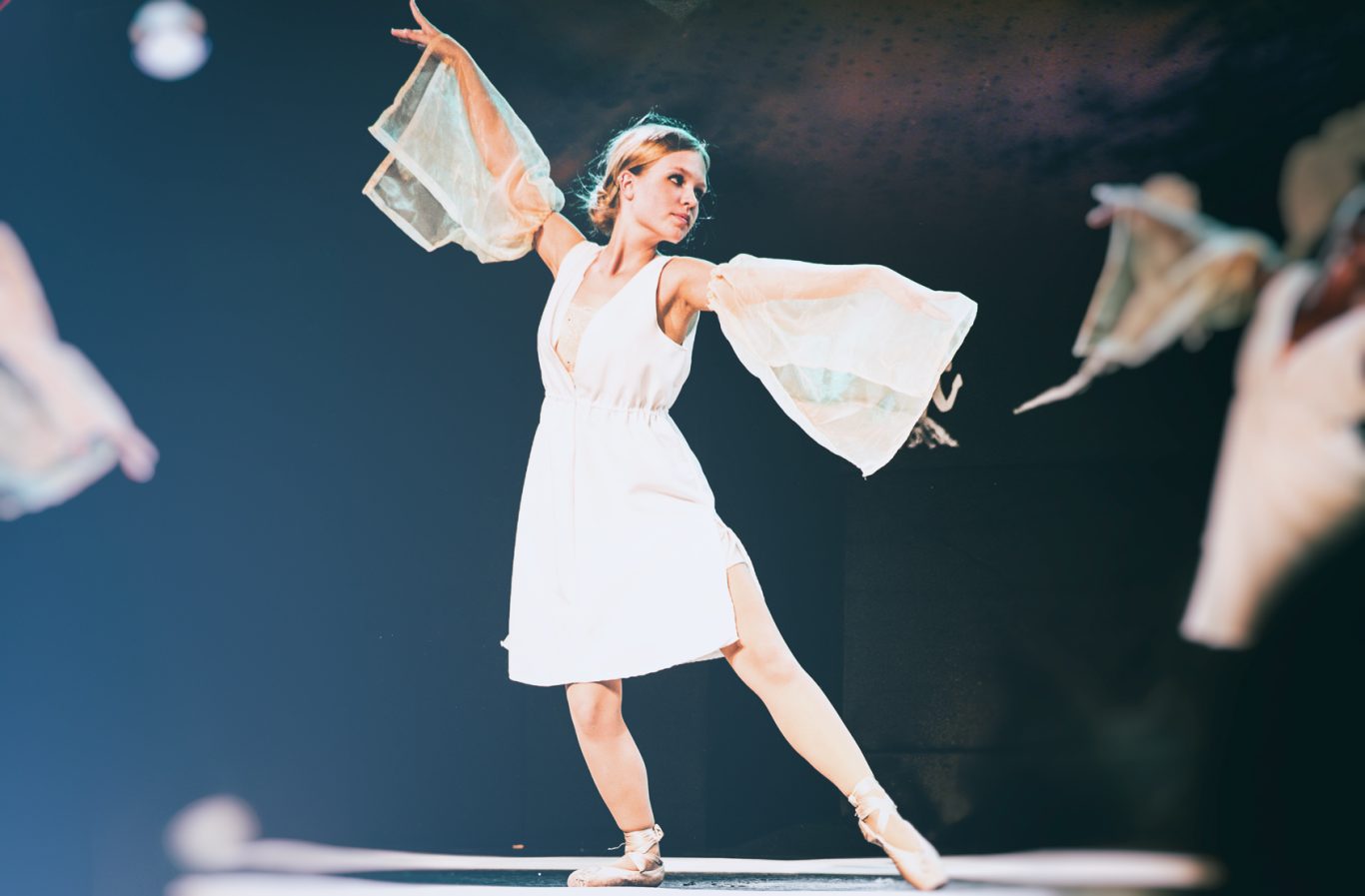 Balletttänzerin in weißem Kleid auf Bühne, Pose mit ausgestrecktem Arm und Bein.