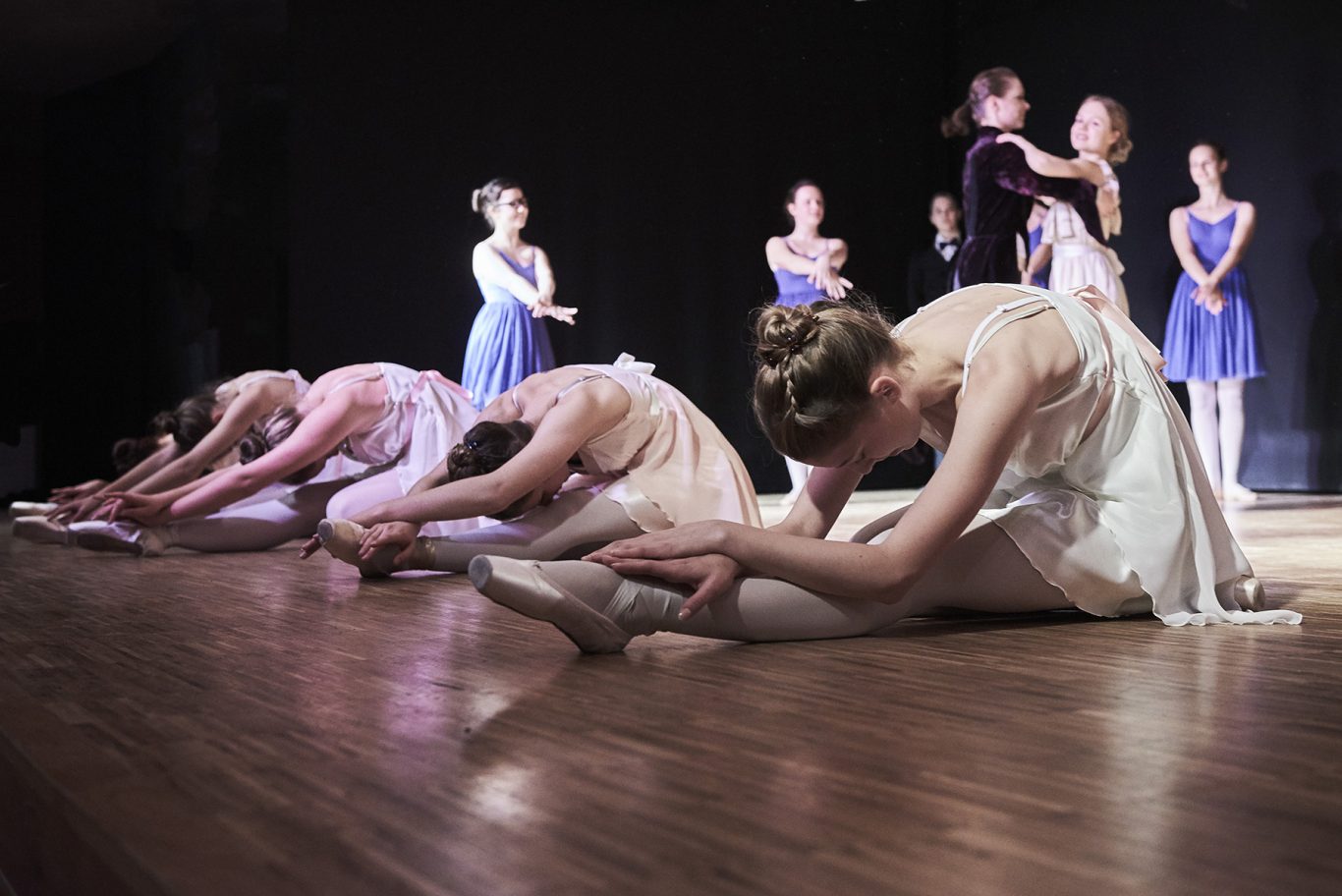 Balletttänzerinnen in weißen Kostümen verbeugen sich auf einer Bühne, im Hintergrund weitere Tänzer*innen.