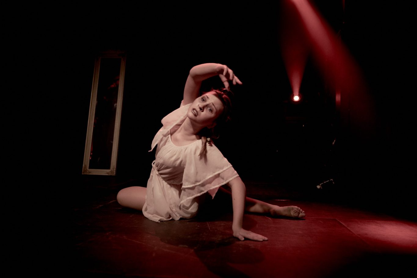 Ballerina in weißem Tutu sitzt auf Bühnenboden, eine Hand am Kopf, rotes Bühnenlicht.